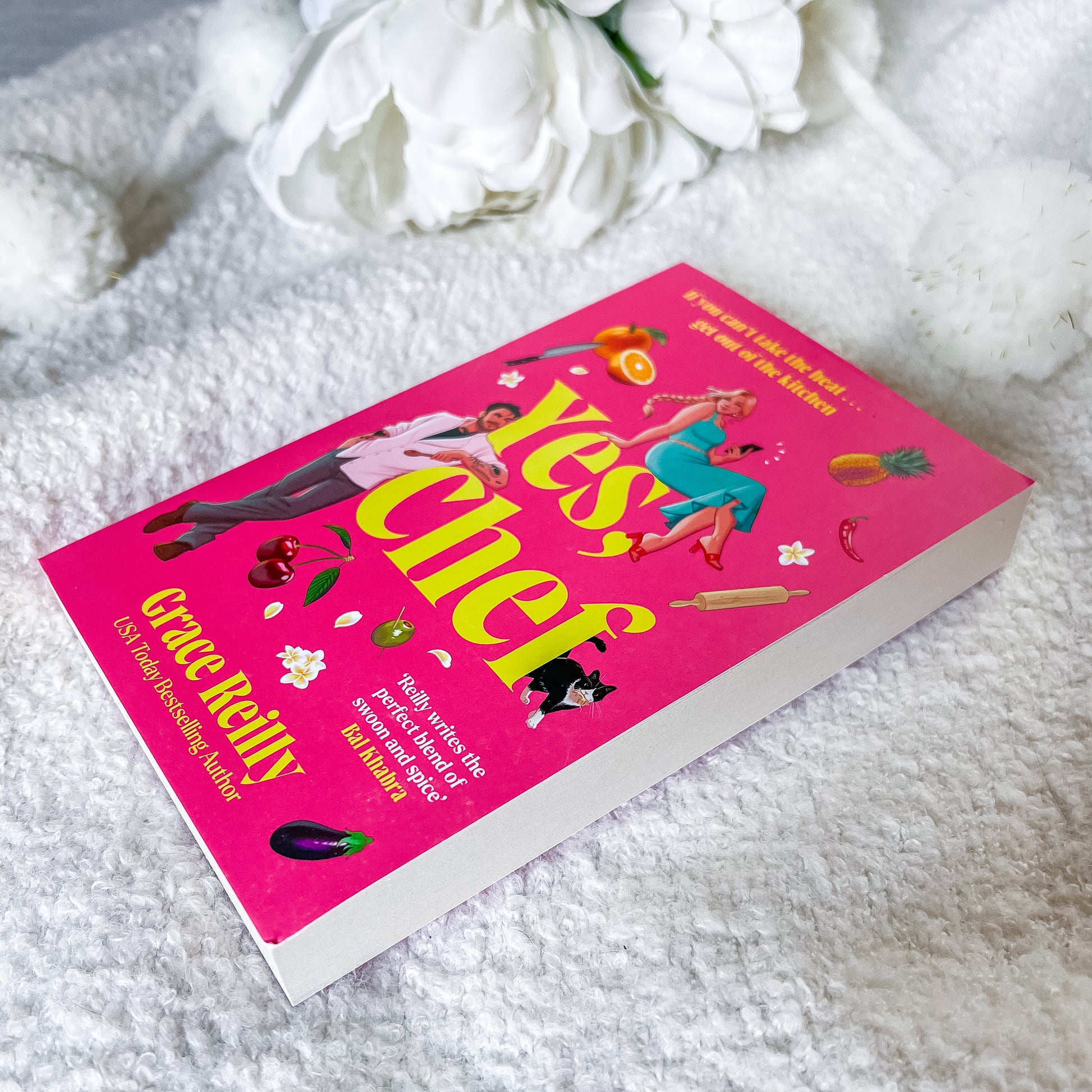 A hardcover book titled 'Yes, Chef' by Grace Kelly, featuring a bright pink cover with large yellow title text and colorful food illustrations, photographed on a white textured blanket with white flowers in the background.