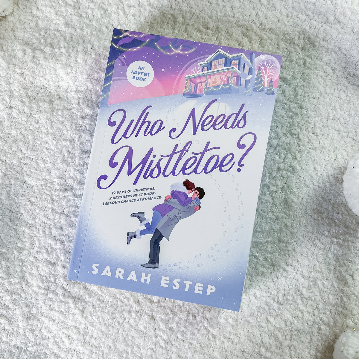 A paperback book titled "Who Needs Mistletoe?" by Sarah Estep, featuring a festive Christmas-themed cover illustration, placed on a soft white textured blanket.