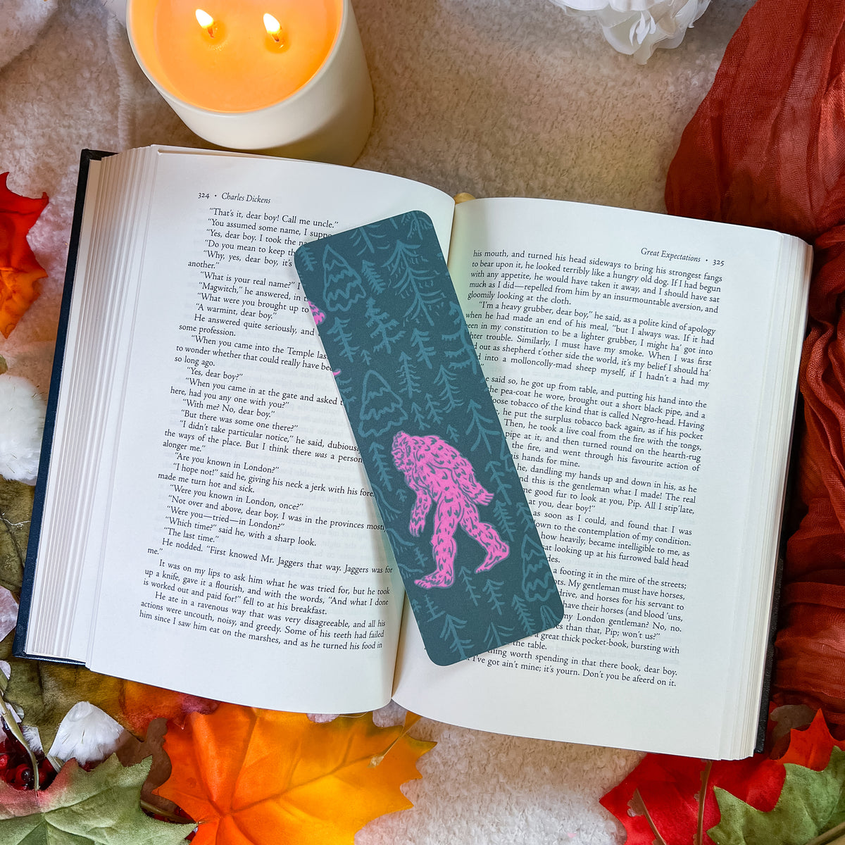 A dark teal bookmark featuring a pink Bigfoot illustration and pine tree pattern, resting on an open book surrounded by autumn leaves, a lit candle, and a white flower.
