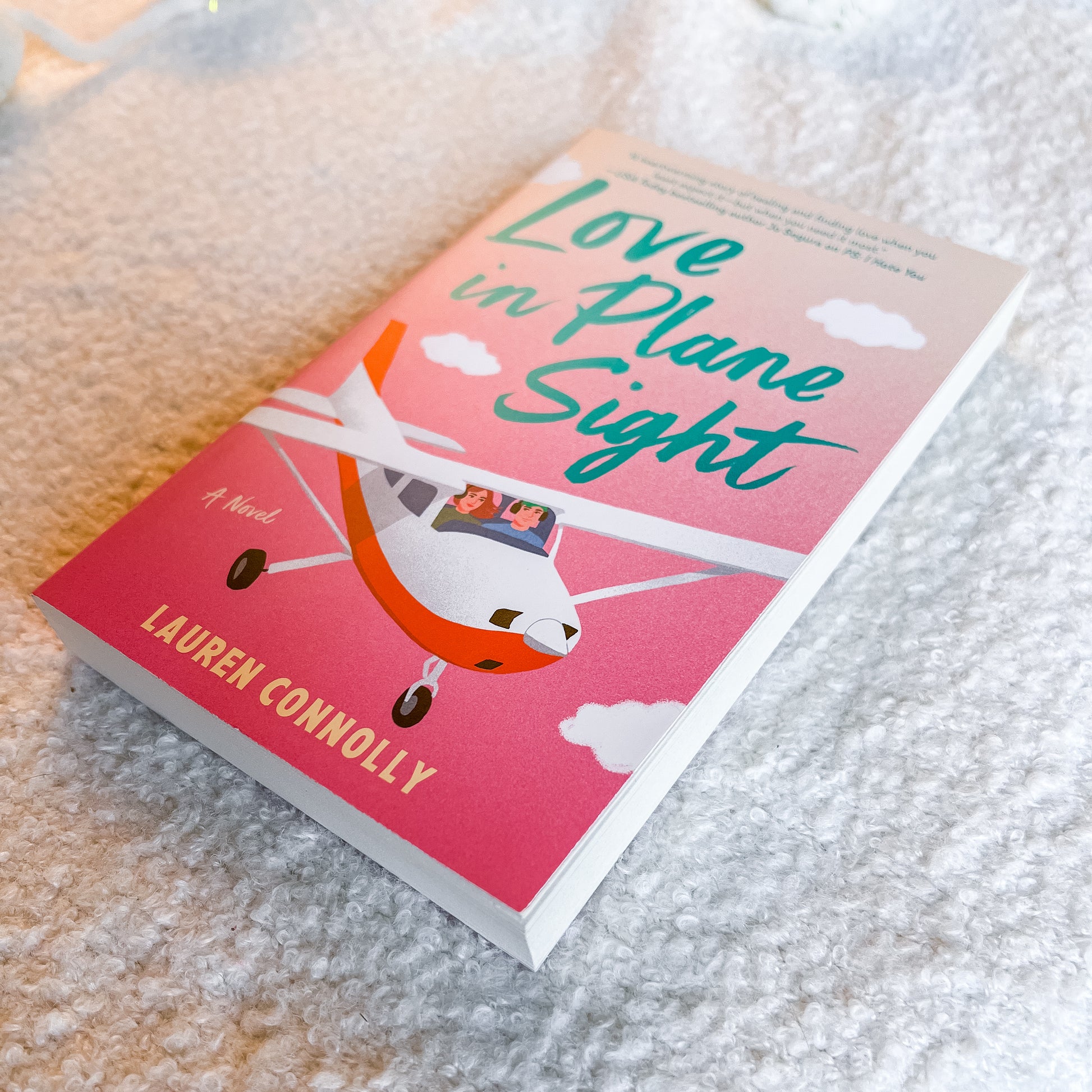 A paperback book titled 'Love in Plane Sight' by Lauren Connolly, featuring a pink and orange cover with an illustration of a small airplane, placed on a textured white blanket.