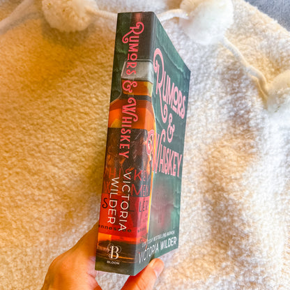 Book titled 'Rumors & Whiskey' held by a hand on a textured surface