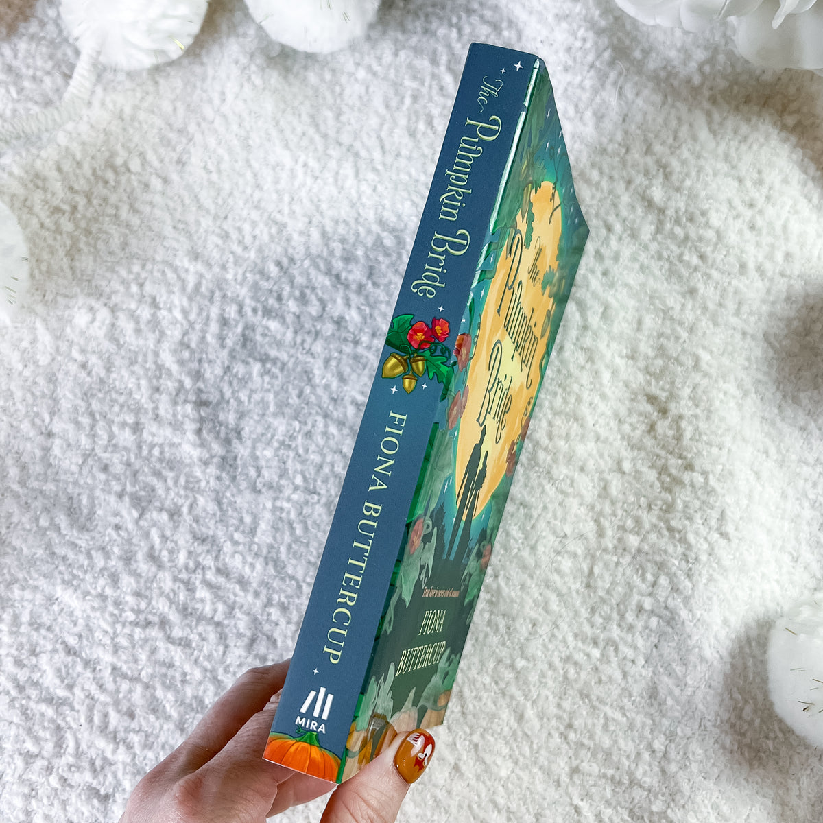 A hand holds a copy of the book by Fiona Buttercup, showing the blue spine with gold lettering and colorful cover art, set against a soft white textured blanket background.
