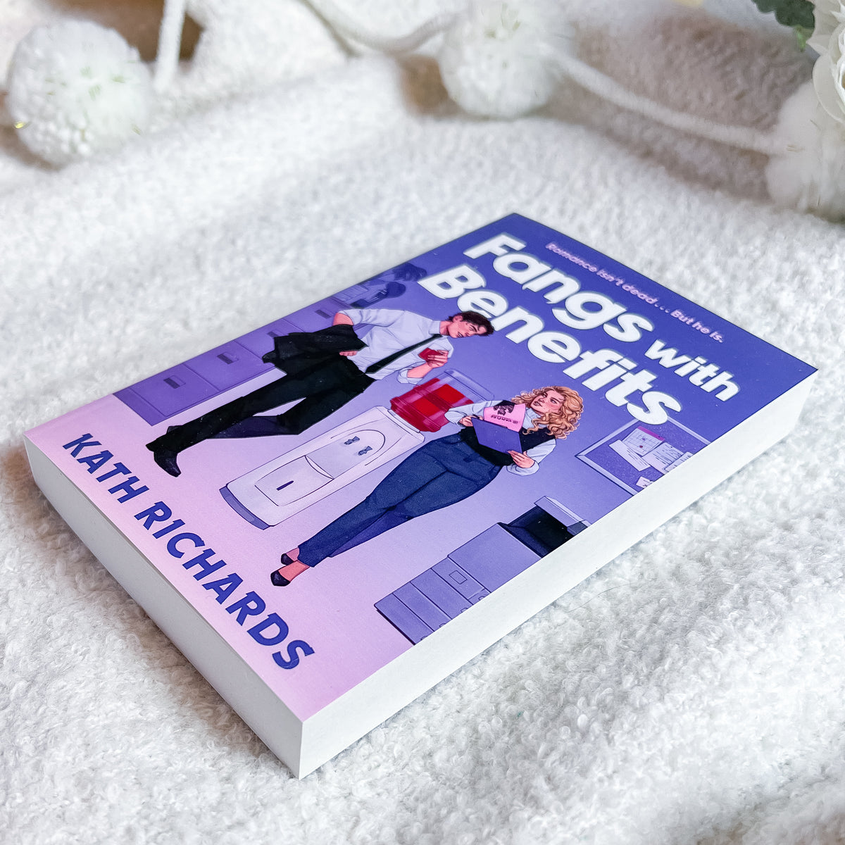 A paperback book titled "Fangs with Benefits" by Kath Richards, featuring a purple and pink cover with an illustration of two office workers, photographed on a soft white textured blanket.