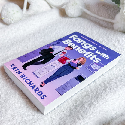 A paperback book titled "Fangs with Benefits" by Kath Richards, featuring a purple and pink cover with an illustration of two office workers, photographed on a soft white textured blanket.