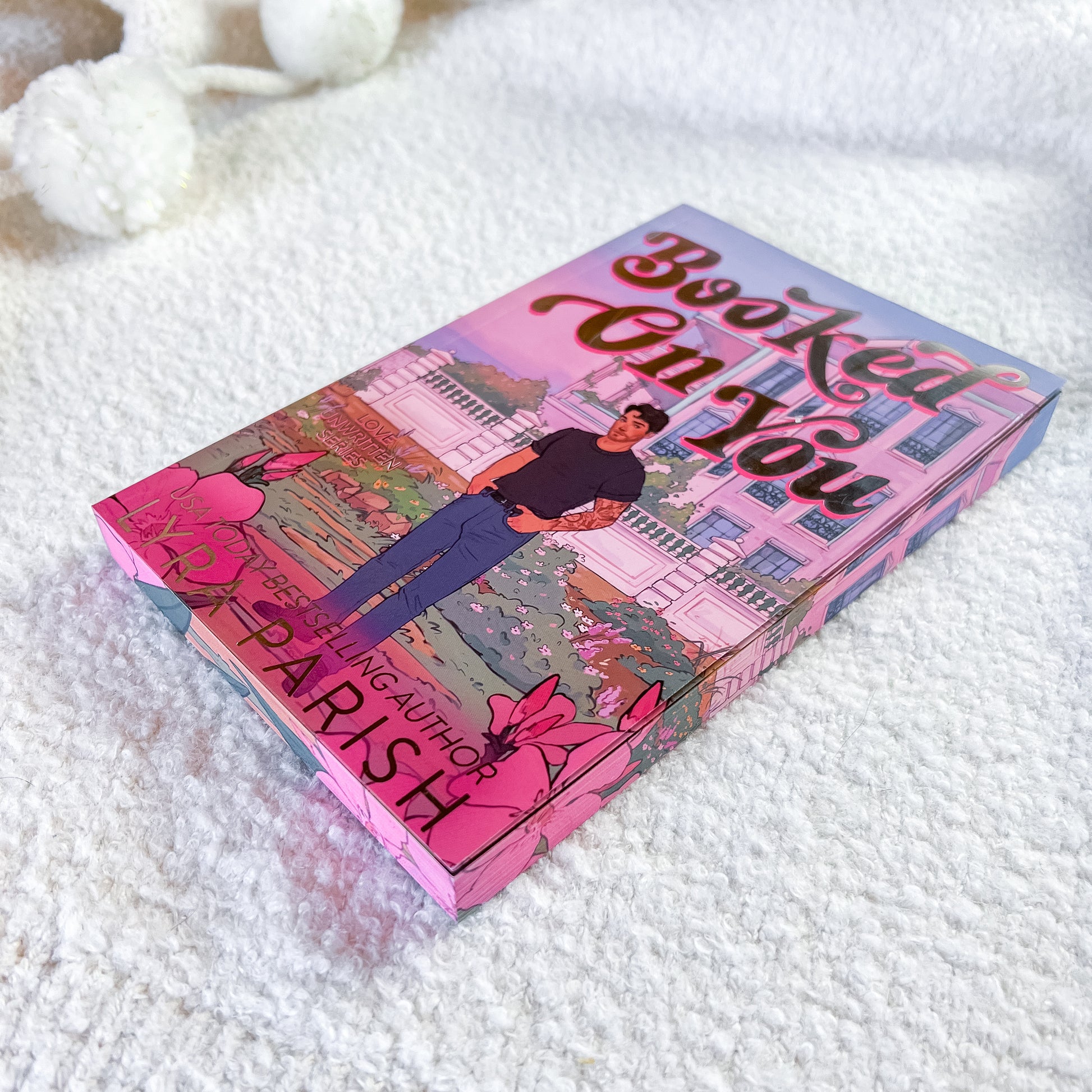 A book titled 'Booked on You' by bestselling author Lyra Parish, featuring a colorful cover illustration of a man standing in front of a pink mansion, placed on a soft white textured blanket.