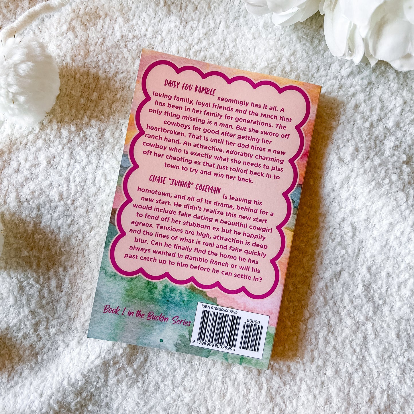 The back cover of a book is displayed flat on a white textured blanket with white flowers in the background. The cover features a pink wavy border around a light peach background with black text, and a small illustration of a cowboy boot and horseshoe at the bottom left.