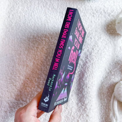 A hand holds a book. The book cover is dark purple with pink and white text and illustrations, set against a soft white textured blanket background.