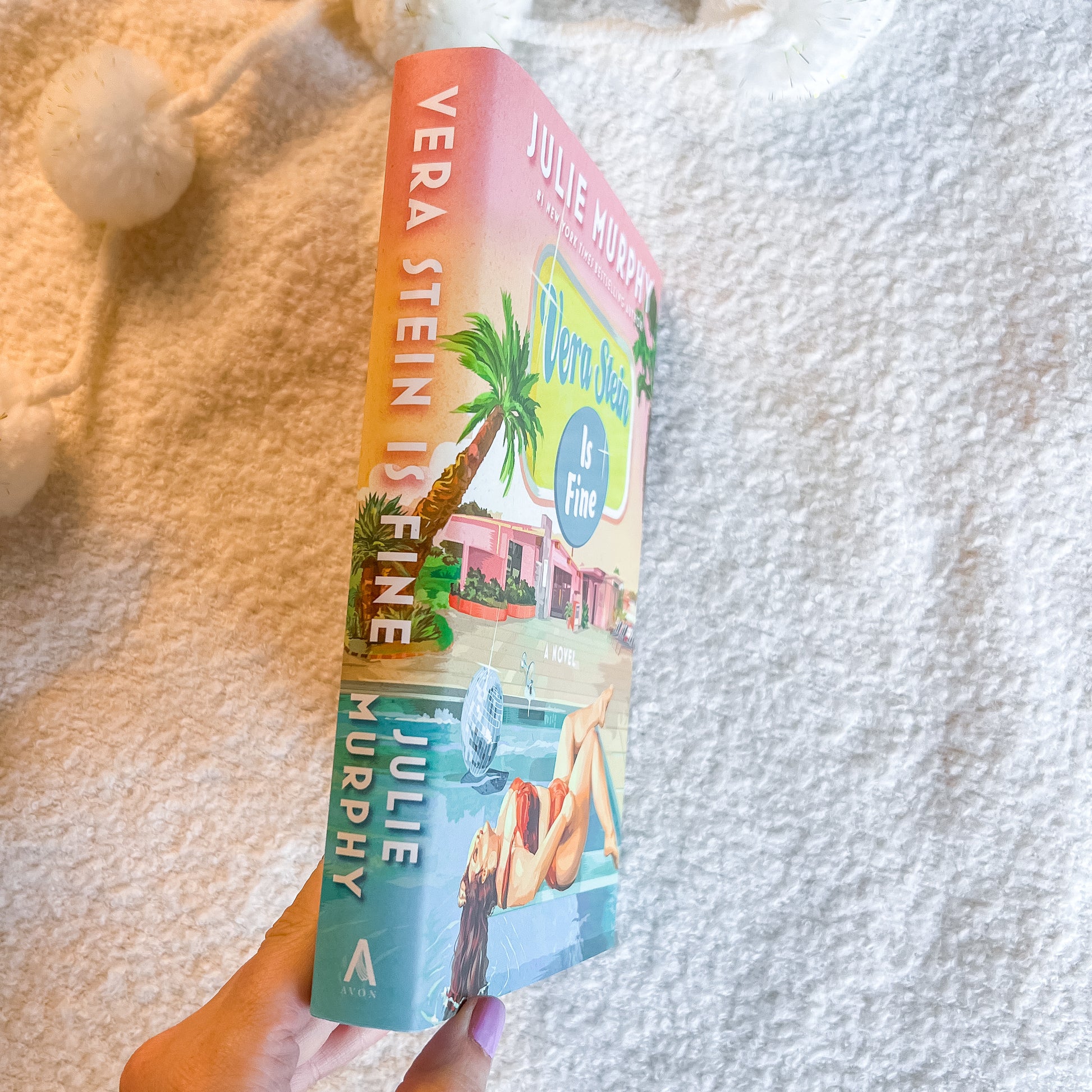 A hand holds a copy of the book by Julie Murphy, showing the colorful cover art with a pink beach house and palm tree against a textured white blanket background.
