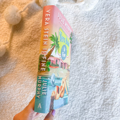A hand holds a copy of the book by Julie Murphy, showing the colorful cover art with a pink beach house and palm tree against a textured white blanket background.