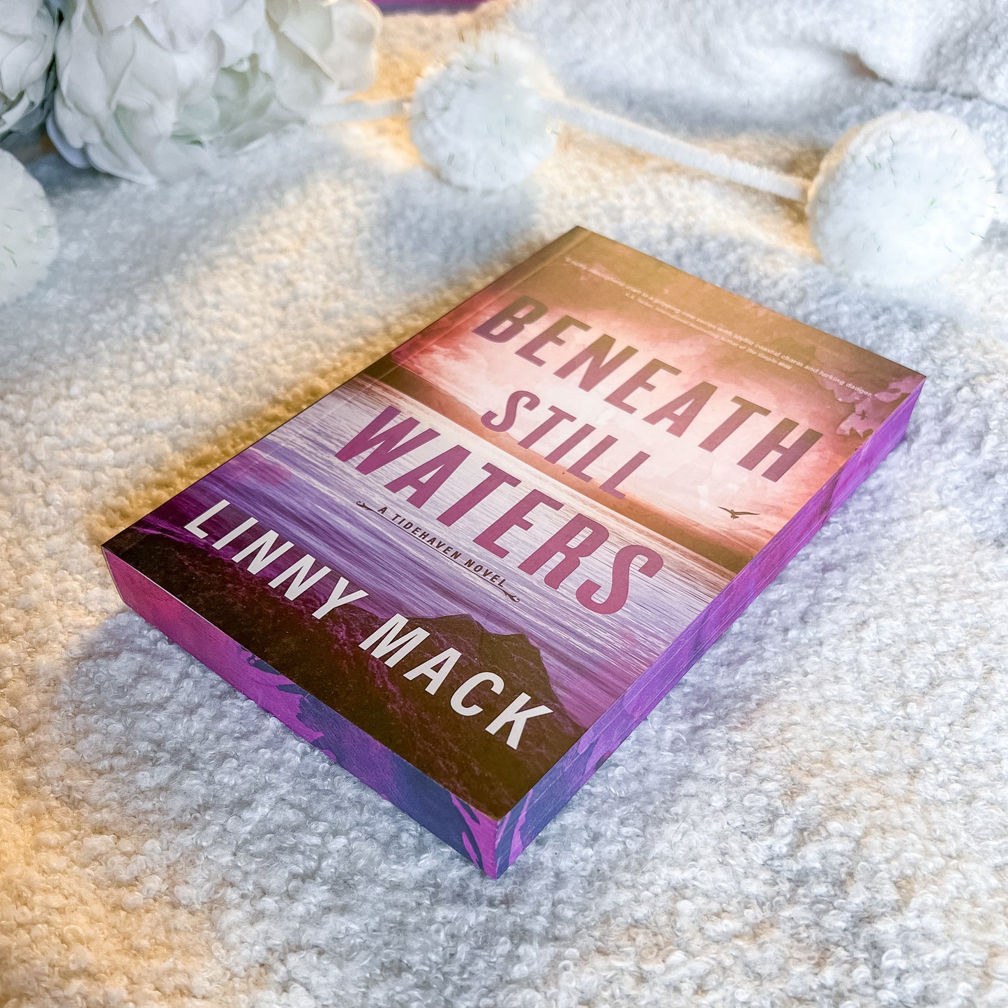Book titled 'Beneath Still Waters' by Linny Mack on a textured surface with flowers.
