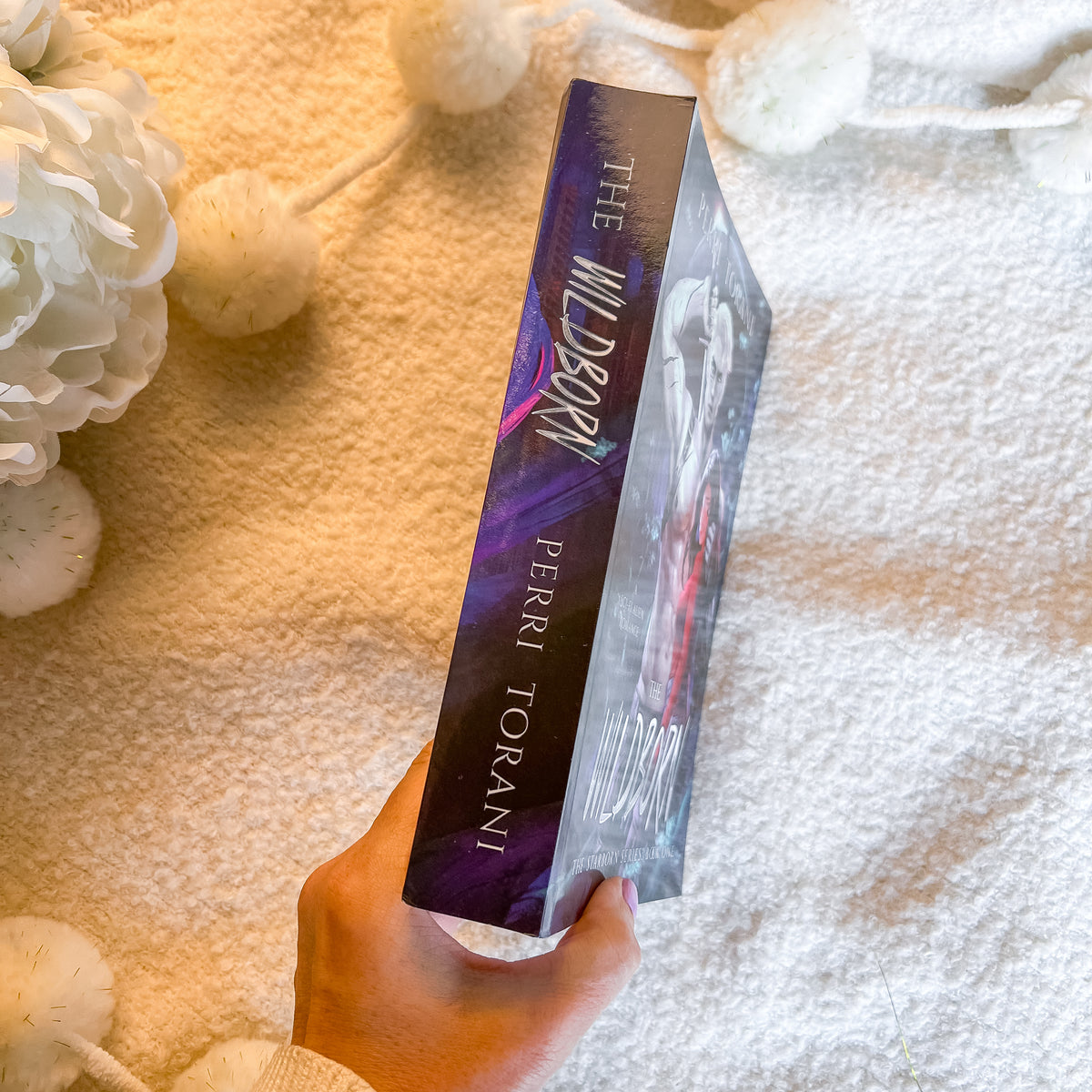 A hand holds a copy of the book 'The Wildbook' by Perri Torani, showing the purple and black spine with white lettering, set against a cozy white textured blanket with decorative white flowers.
