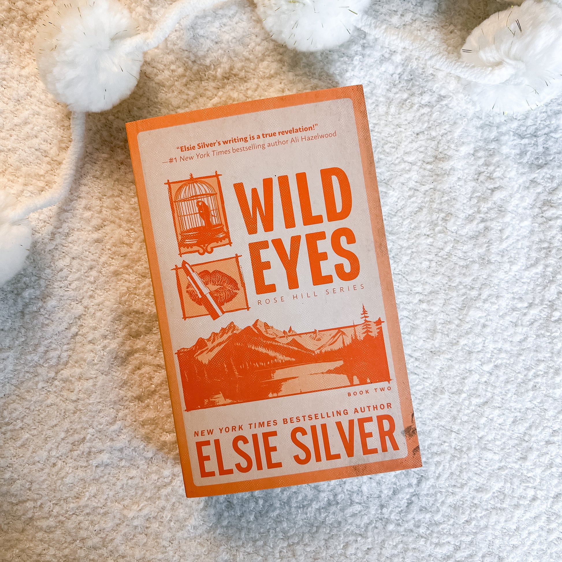 Book cover for 'Wild Eyes' by Elsie Silver, featuring orange and cream artwork with illustrations of a birdcage, pen, and mountains, photographed on a textured white blanket with white pom-pom yarn in the background.