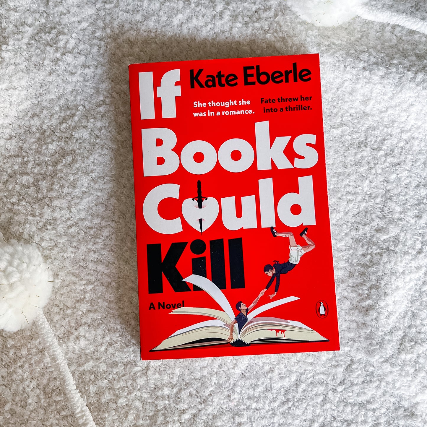 The book cover of 'If Books Could Kill' by Kate Eberle is displayed flat on a textured white blanket. The cover is predominantly bright red with bold white and black typography, featuring an illustration of a person falling off an open book.