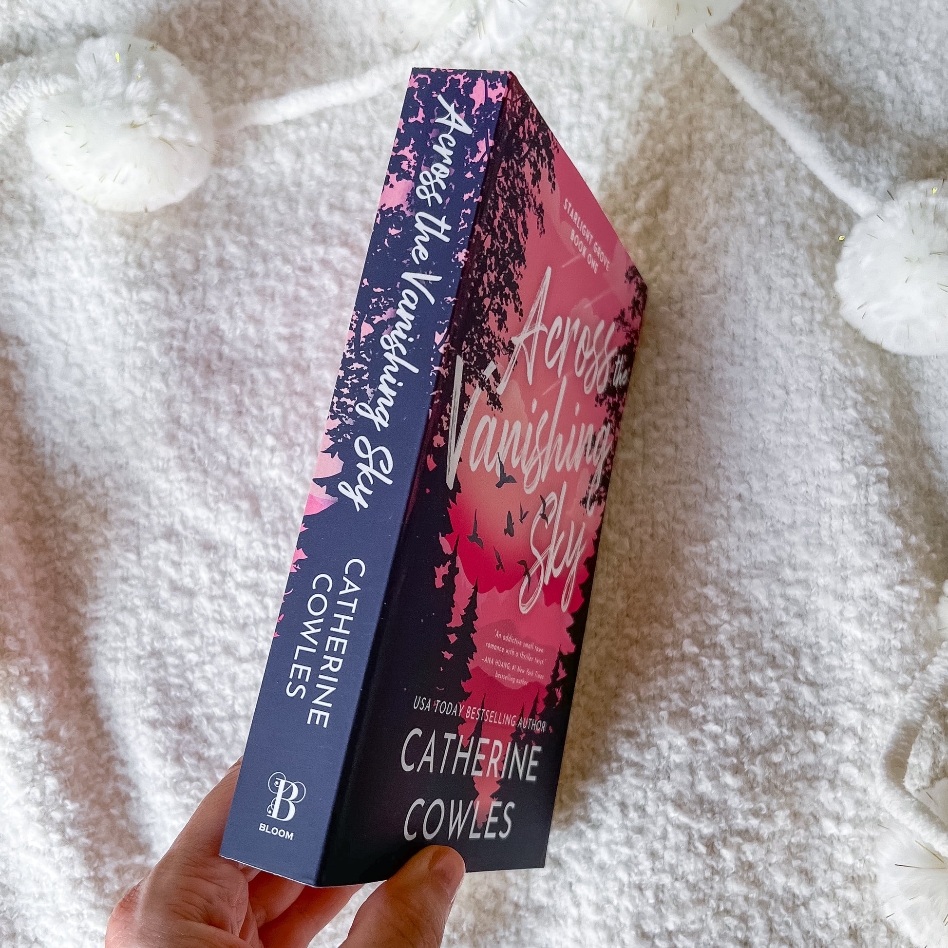 A hand holds a paperback copy of the book 'Across a Vanishing Sky' by Catherine Cowles, showing both the dark navy spine and the pink-to-purple illustrated front cover, set against a soft white textured blanket background.