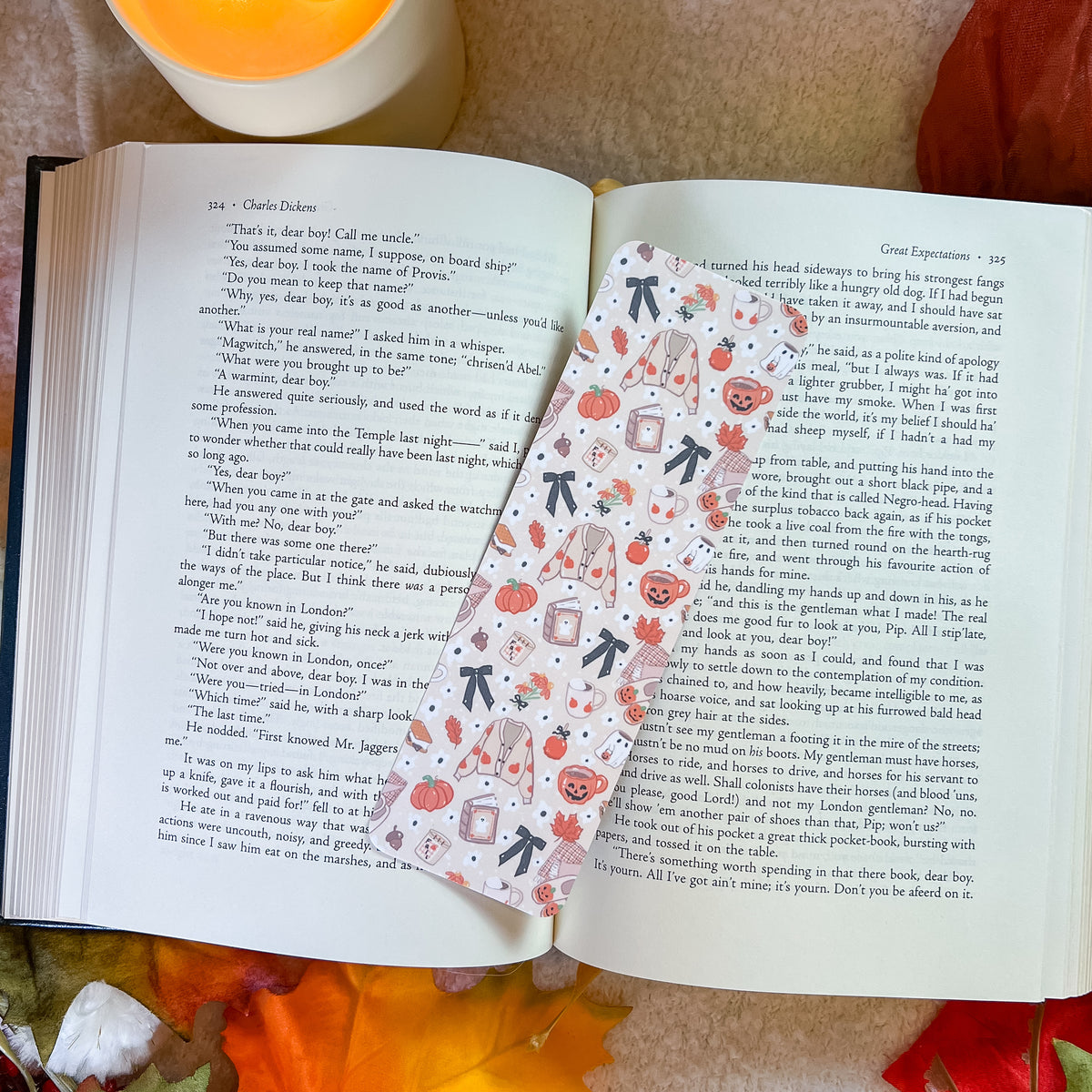 A decorative paper bookmark with a pumpkin and tea cup pattern is shown resting on open pages of a book, with a warm orange blanket in the background.