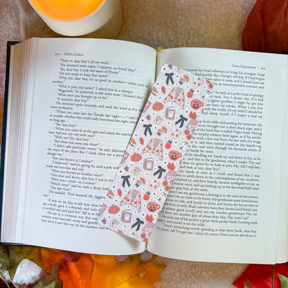 A decorative paper bookmark with a pumpkin and tea cup pattern is shown resting on open pages of a book, with a warm orange blanket in the background.