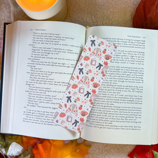 A decorative paper bookmark with a pumpkin and tea cup pattern is shown resting on open pages of a book, with a warm orange blanket in the background.