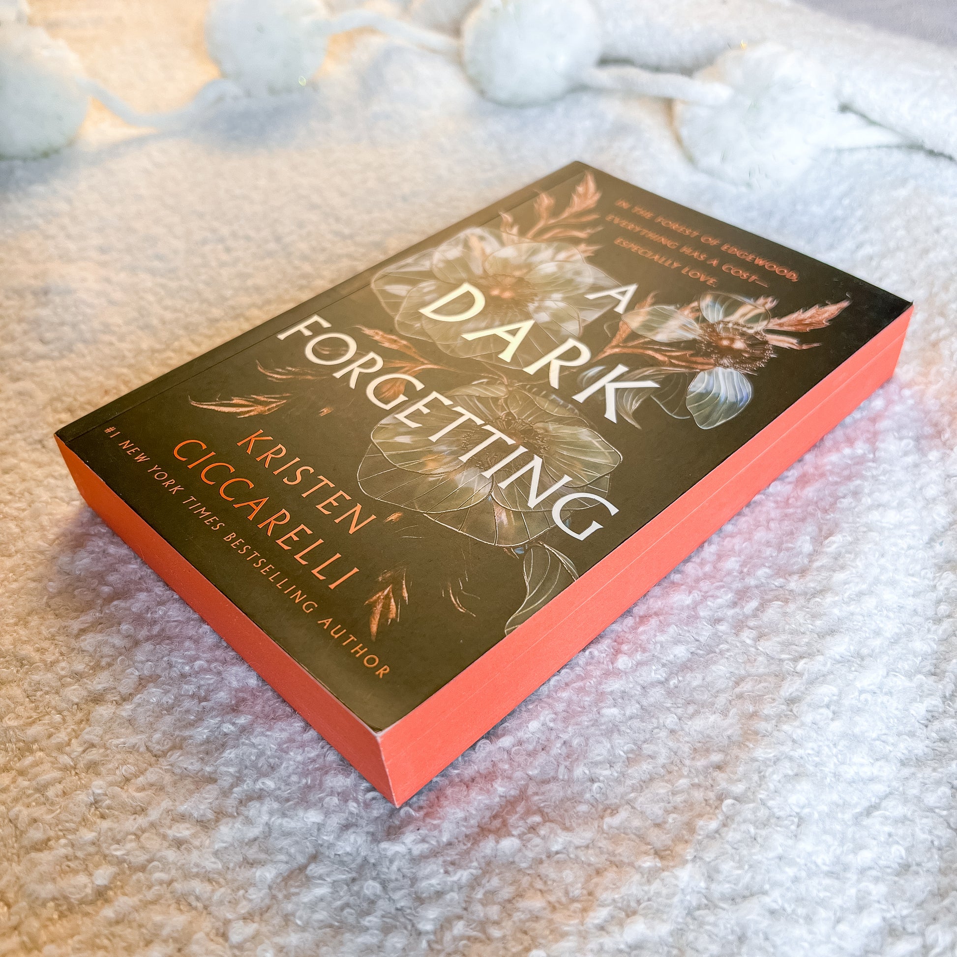 A book titled 'A Dark Forgetting' by Kristen Giccarelli, featuring a dark olive-green cover with floral illustrations and a bright coral-orange spine, resting on a textured white blanket.
