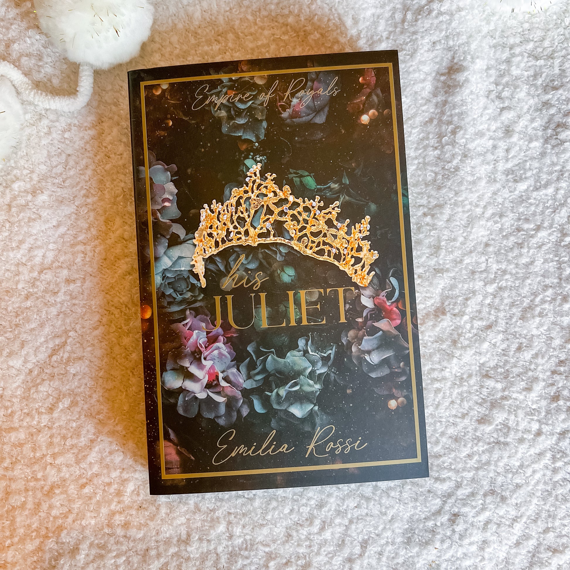 A book titled 'Bis Juliet' by Emilia Rossi, featuring a dark cover with a gold tiara illustration and gold lettering, placed on a soft white textured blanket.
