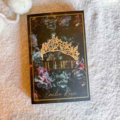 A book titled 'Bis Juliet' by Emilia Rossi, featuring a dark cover with a gold tiara illustration and gold lettering, placed on a soft white textured blanket.
