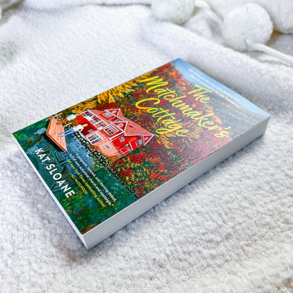 A book by Kat Sloane, featuring a colorful cover illustration of a red cottage in a flower-filled field, photographed on a soft white textured blanket.