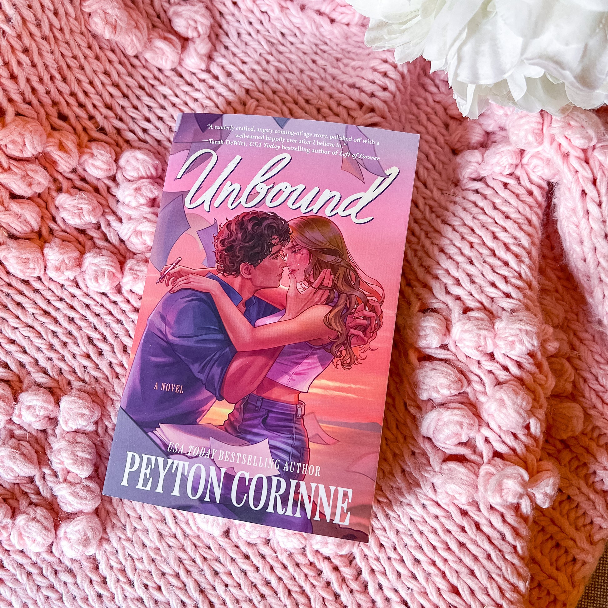 Book titled 'Unbound' by Peyton Corinne on a pink textured surface with white flowers.