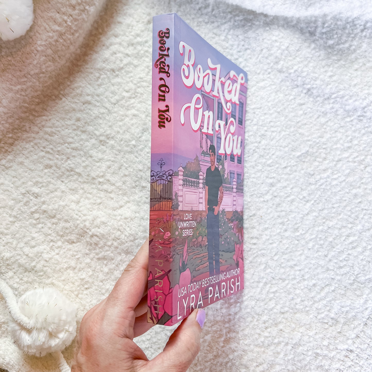 A hand holds a paperback copy of the book 'Booked Up You' by Lyra Parish against a soft white textured blanket background. The book cover features a pink and purple-toned illustration of a woman standing in front of a building.