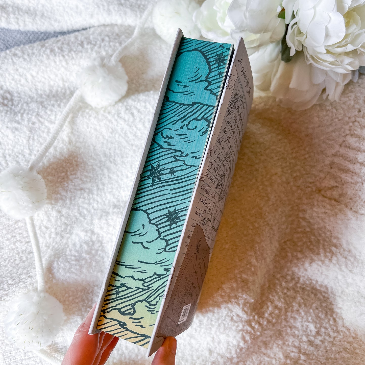 Book with decorative cover held by a hand on a textured surface with flowers in the background