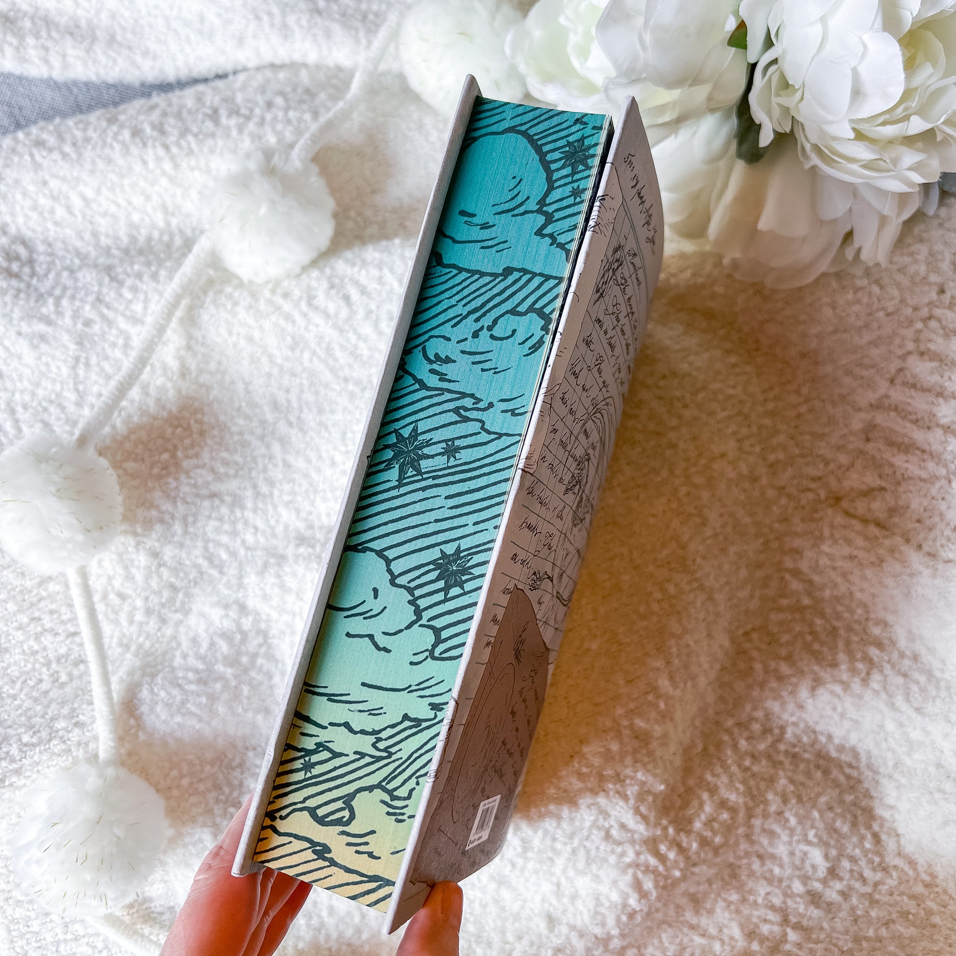 Book with decorative cover held by a hand on a textured surface with flowers in the background