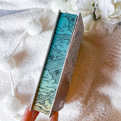 Book with decorative cover held by a hand on a textured surface with flowers in the background