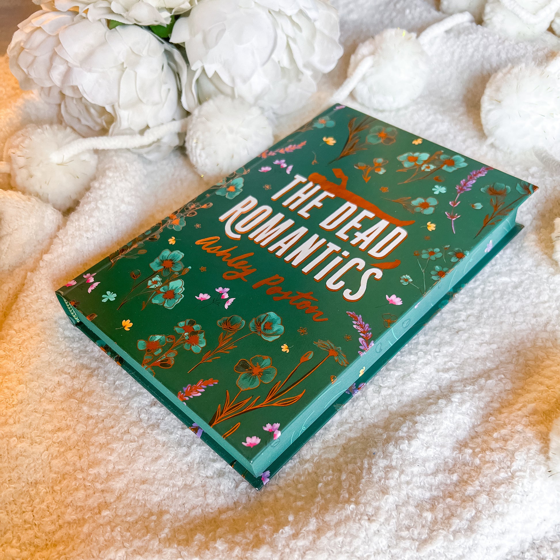 Book titled 'The Dead Romantics' by Ruby Pastor on a soft surface with flowers.