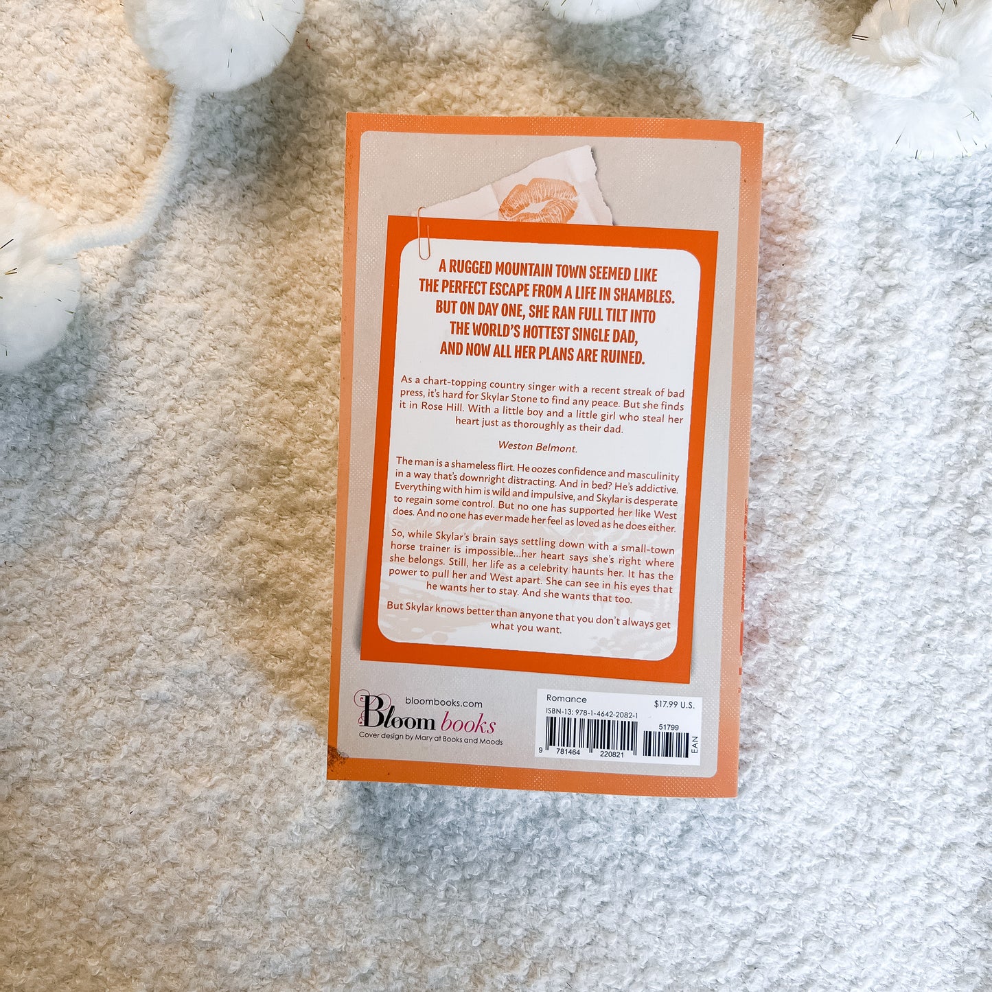 The back cover of a book by Bloom Books, featuring an orange border and white background with black text, photographed flat on a white textured blanket.