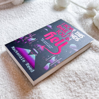 A hardcover book titled 'I Hope This Email Finds You In Hell' by Mackenzie Reed, featuring a dark cover with pink and purple cosmic illustrations, resting on a soft white textured blanket.