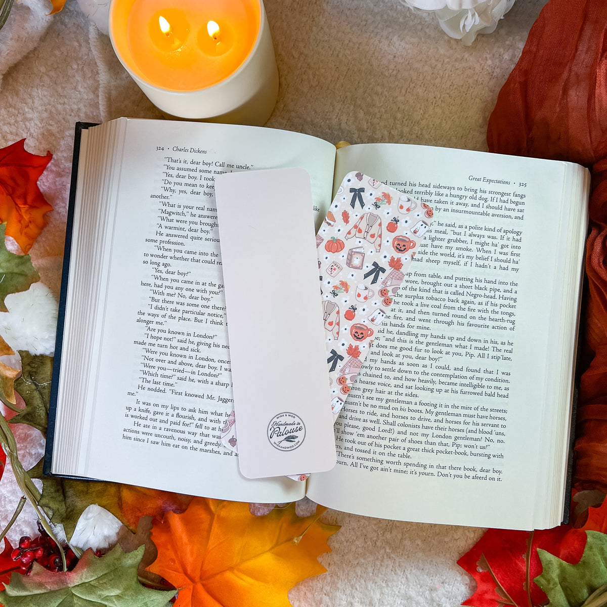 A white rectangular bookmark with a circular logo sits on an open book, alongside a decorative patterned bookmark featuring small illustrations of pumpkins. The scene is styled with autumn leaves, a lit candle, and a cozy blanket background.