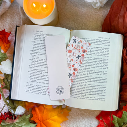 A white rectangular bookmark with a circular logo sits on an open book, alongside a decorative patterned bookmark featuring small illustrations of pumpkins. The scene is styled with autumn leaves, a lit candle, and a cozy blanket background.