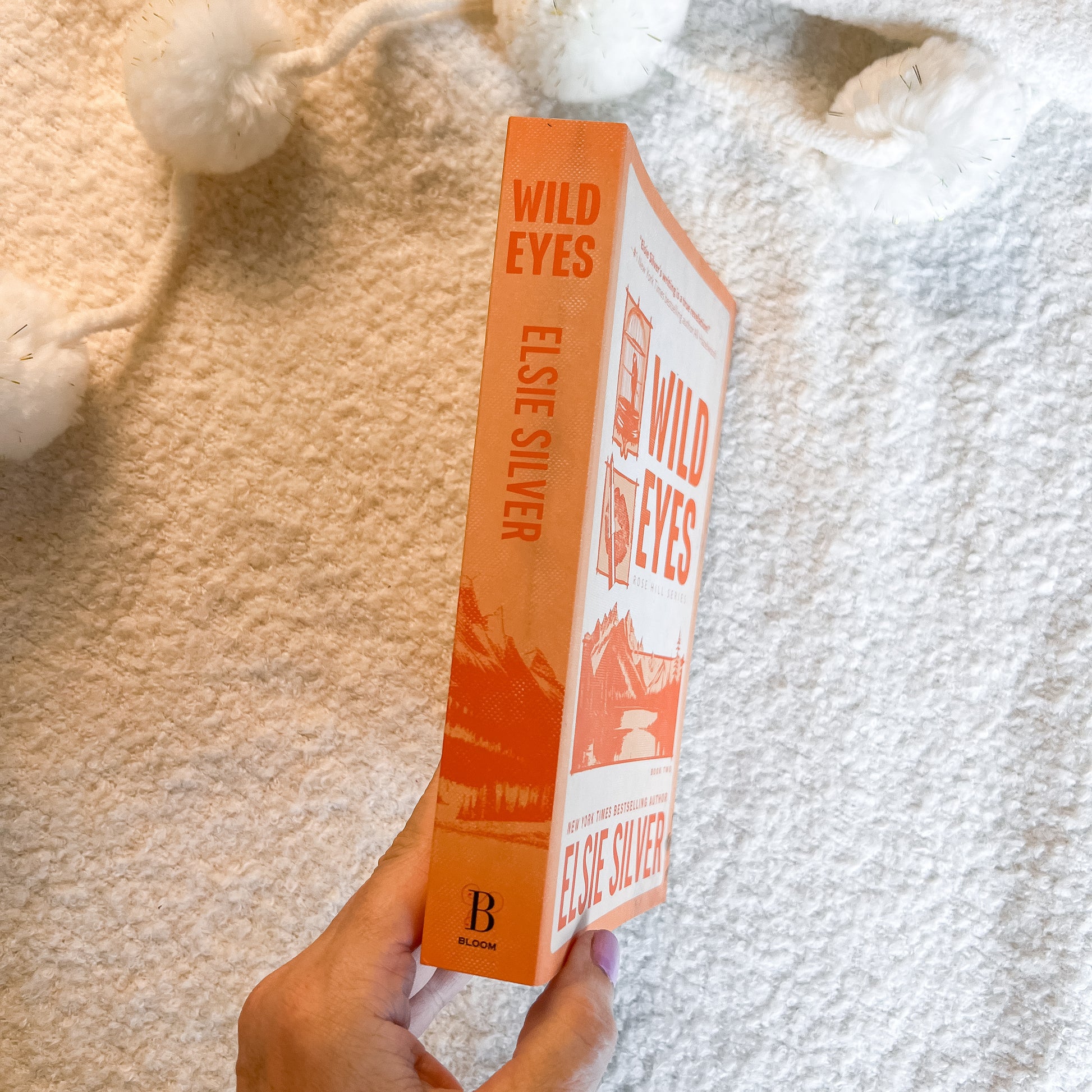 A hand holds a copy of the book 'Wild Eyes' by Elsie Silver, showing the orange and white cover and spine, against a soft beige textured blanket background with white pom-pom tassels.