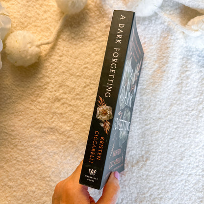 A hand holds a book titled 'A Dark Forgetting' by Kristen Giccarelli, showing the spine with gold floral illustrations and white lettering, set against a soft white textured blanket background.