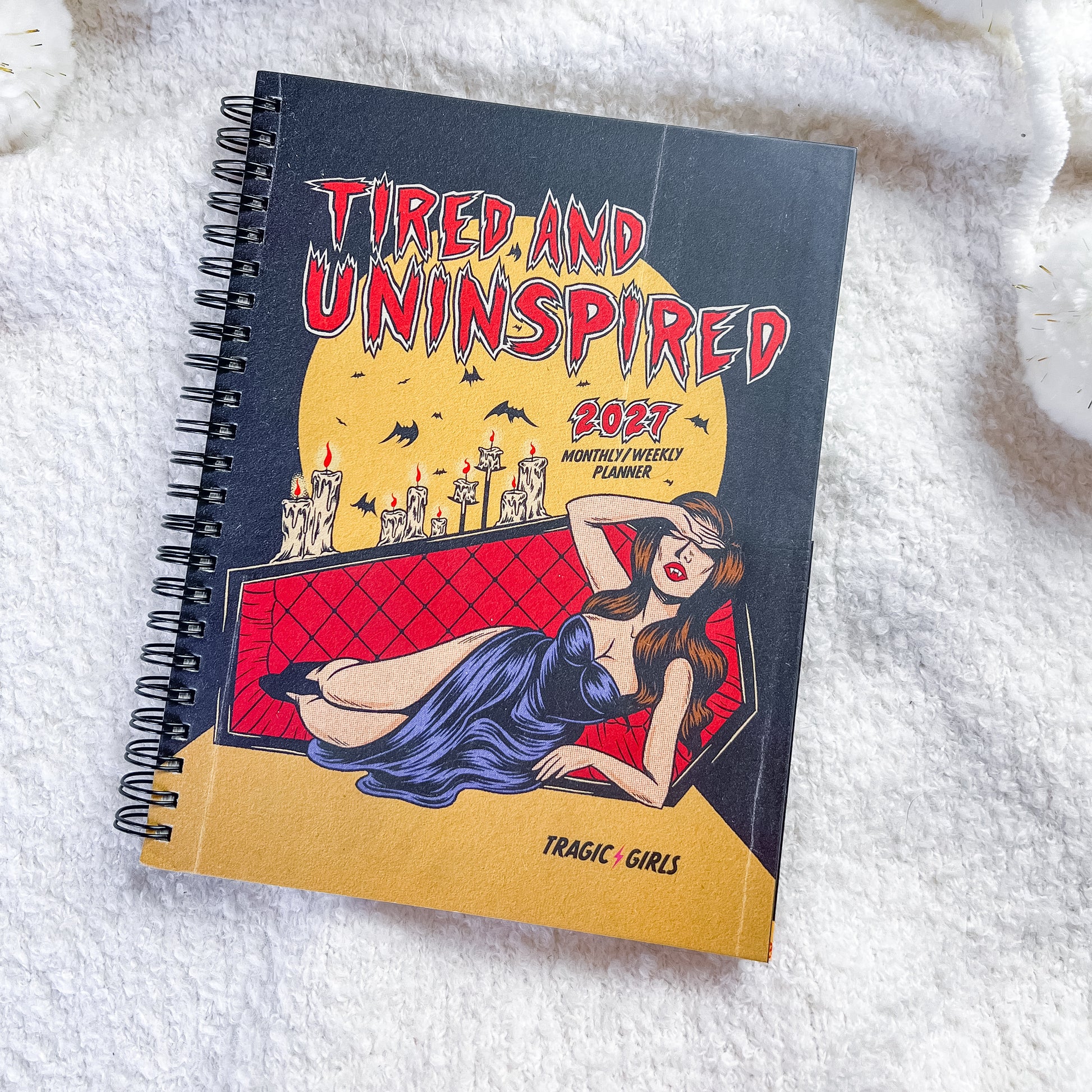 A spiral-bound notebook titled 'TIRED AND UNINSPIRED 2027 MORTAL CORPSE PLANNER' with a dark cover featuring a pin-up illustration of a woman in a blue dress on a red couch, placed on a white textured blanket.