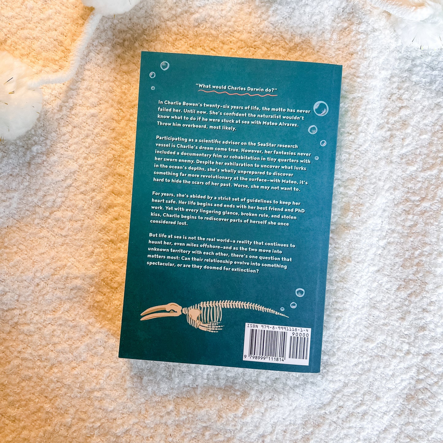 The back cover of a book lying flat on a textured white blanket. The cover is dark teal with white text, a small illustration of a shark skeleton, and a barcode in the bottom right corner.