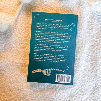 The back cover of a book lying flat on a textured white blanket. The cover is dark teal with white text, a small illustration of a shark skeleton, and a barcode in the bottom right corner.