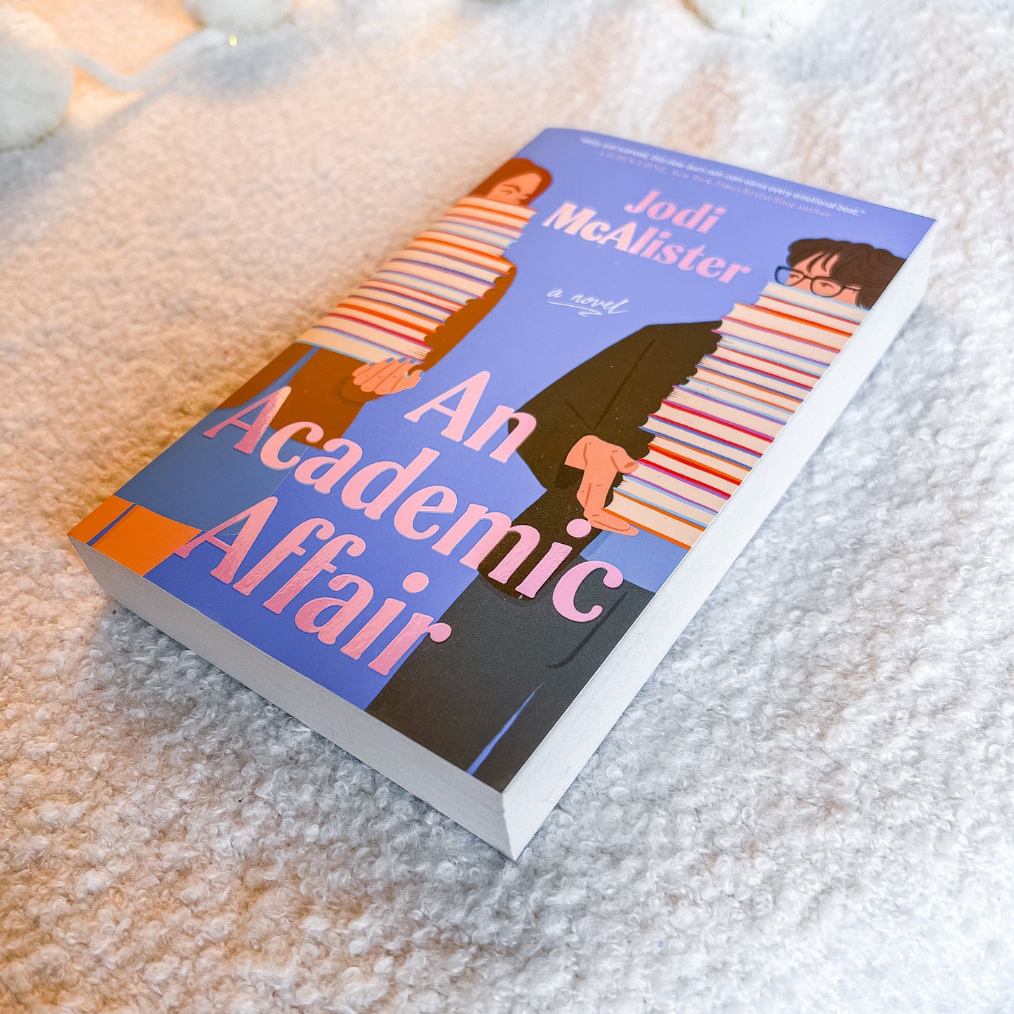 The book 'An Academic Affair' by Jodi McAlister lies on a soft, textured white blanket. The cover features a blue background with an illustration of two students in striped sweaters and a black blazer, with the title in bold pink text.