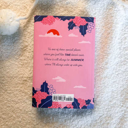 A pink book or notebook lying on a soft beige textured blanket, showing the back cover with floral illustrations in pink and navy, a red sun, white clouds, and a handwritten quote.