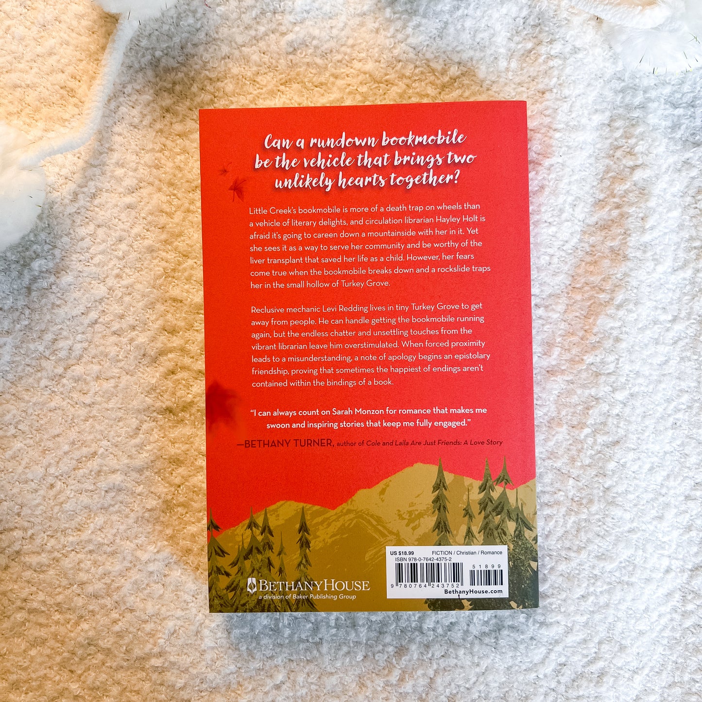 The back cover of the book, featuring a red background with white text and a green forest illustration at the bottom, photographed on a textured beige fabric background.
