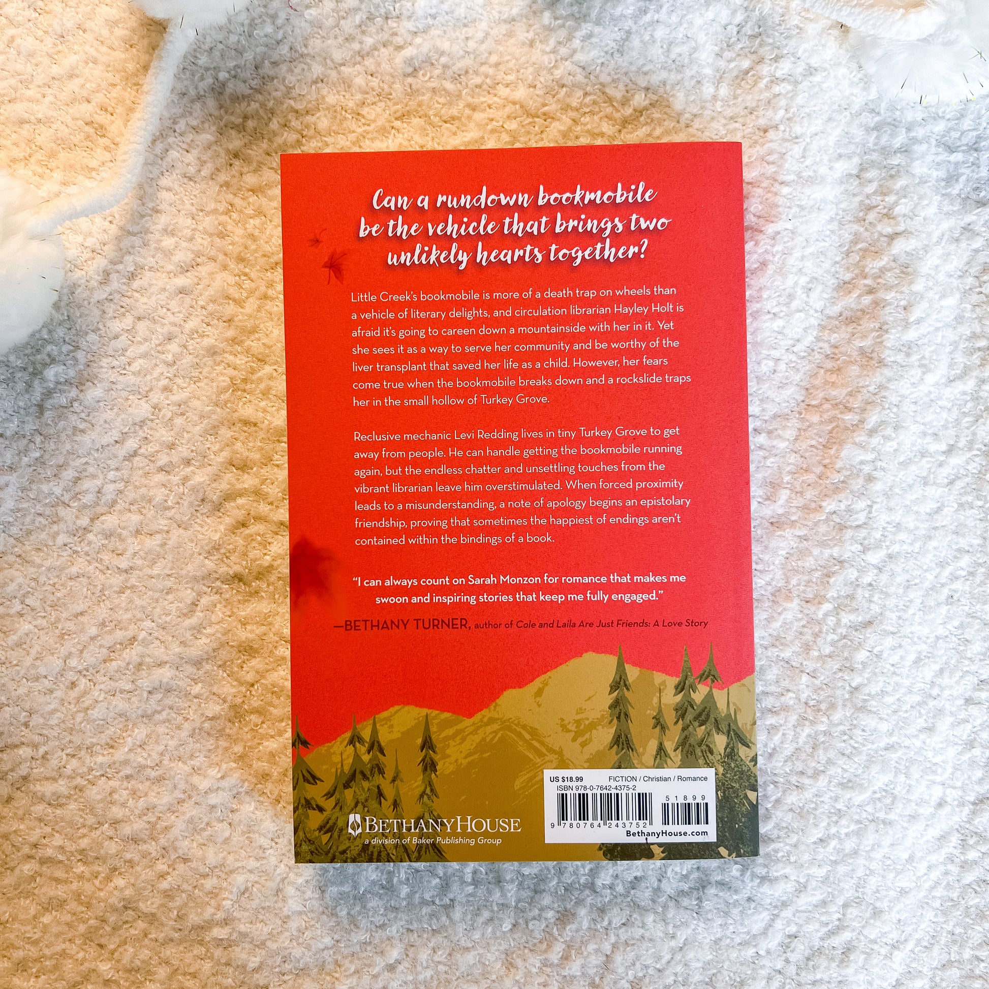 The back cover of the book, featuring a red background with white text and a green forest illustration at the bottom, photographed on a textured beige fabric background.
