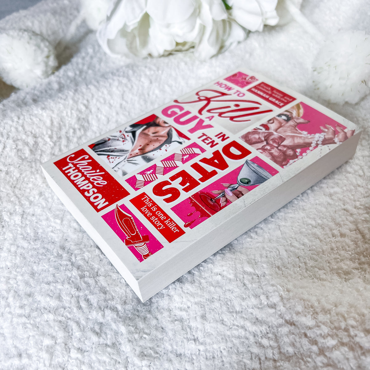 A paperback copy of the book 'How to Kill Guy in Ten Dates' by Shaillee Thompson lies on a white textured blanket with white flowers in the background. The book cover features a vibrant pink and red collage design with various illustrations and text.