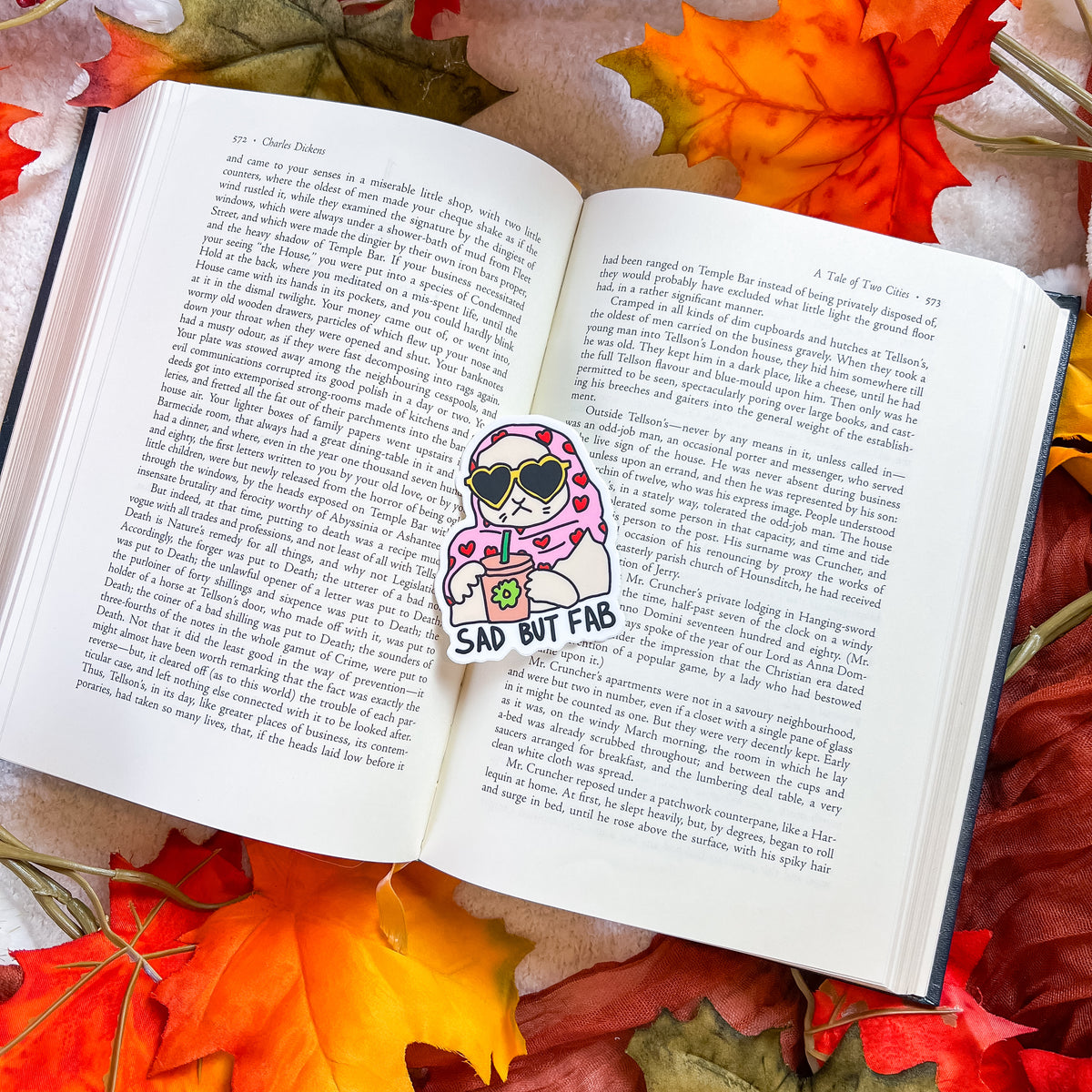 A die-cut sticker featuring a cartoon character with pink hair, sunglasses, and a drink, placed on an open book surrounded by autumn leaves. The sticker has the text 'SAD BUT FAB' printed on it.