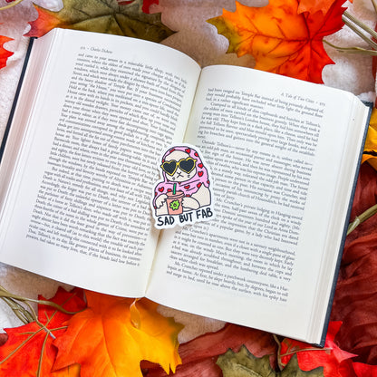 A die-cut sticker featuring a cartoon character with pink hair, sunglasses, and a drink, placed on an open book surrounded by autumn leaves. The sticker has the text 'SAD BUT FAB' printed on it.