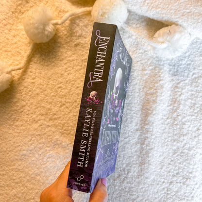 A hand holds a paperback copy of the book by Kayle Smith, showing the purple illustrated cover and spine, set against a cozy white blanket background with white pom-pom garlands.