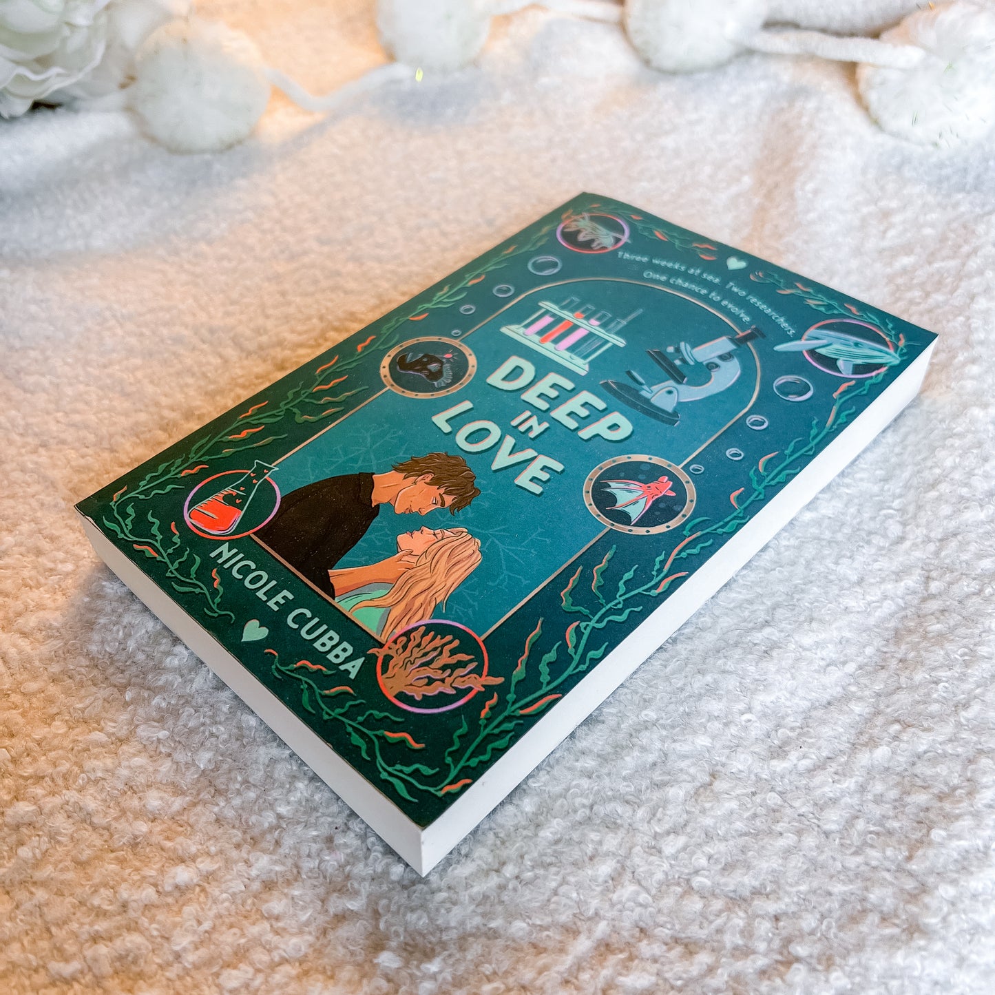 The book 'Deep The Love' by Nicole Cubba, featuring a detailed underwater illustration on its teal cover, resting on a soft, textured cream-colored blanket with white decorative flowers in the background.