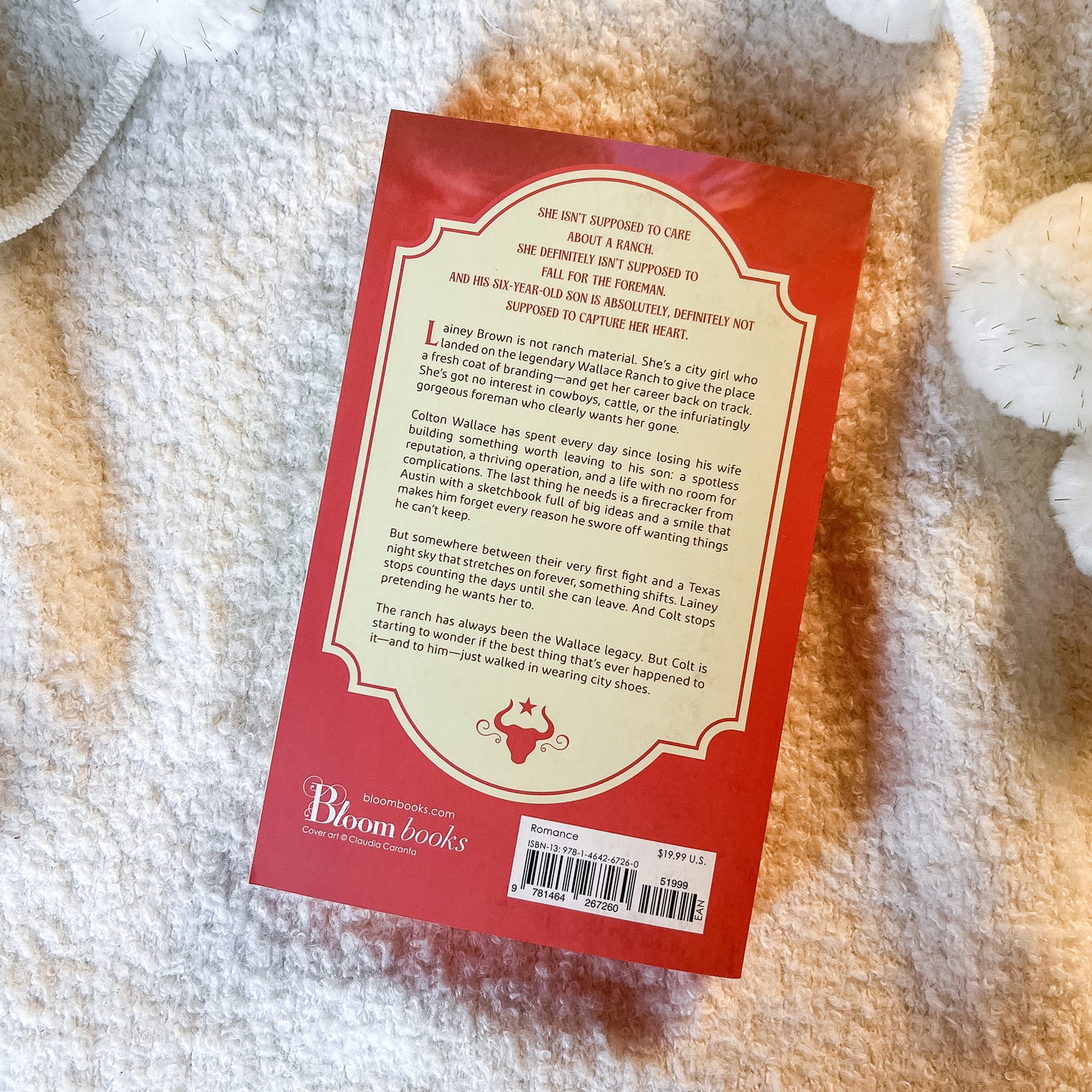 The back cover of a book by Bloom Books, featuring a red border and a cream-colored text block, laid flat on a white textured blanket.